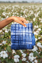 Load image into Gallery viewer, Hand holding a blue checkered tie-dye clutch with a tassel against a cotton field
