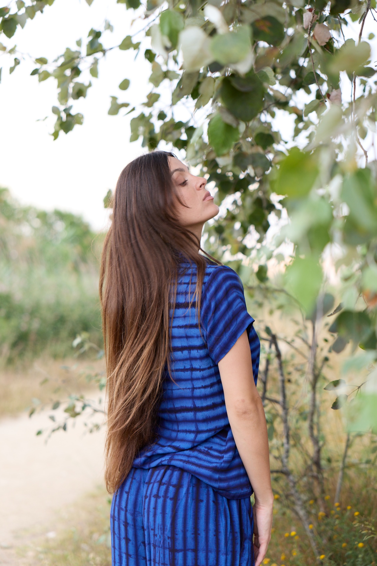 A woman wearing a blue indigo tie & dye outfit standing outdoors among trees.