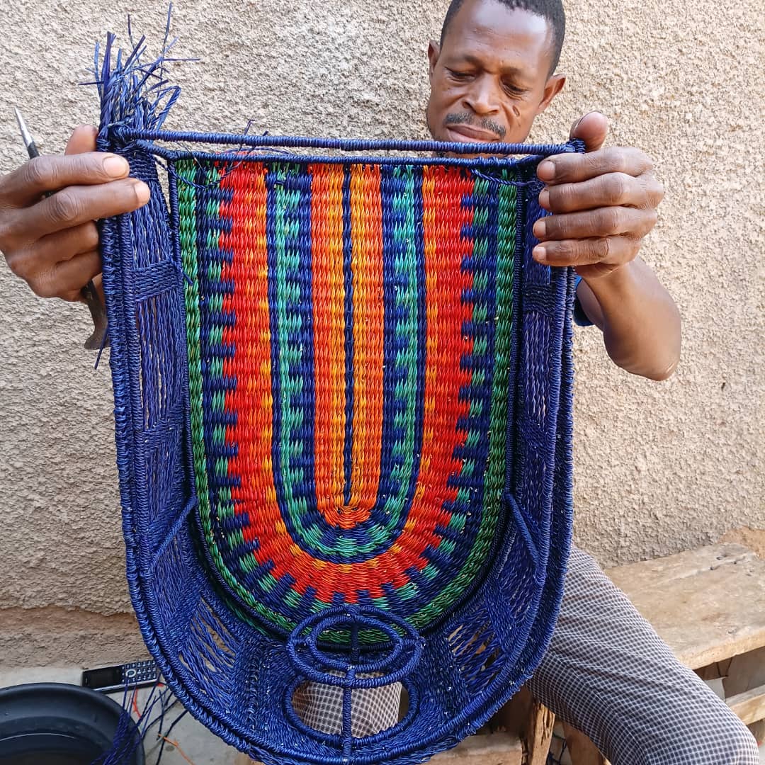 Ghanaian weaver holding the half-finished arch panel of the Amira base, showing the indigo, green, orange and red striped weave in progress