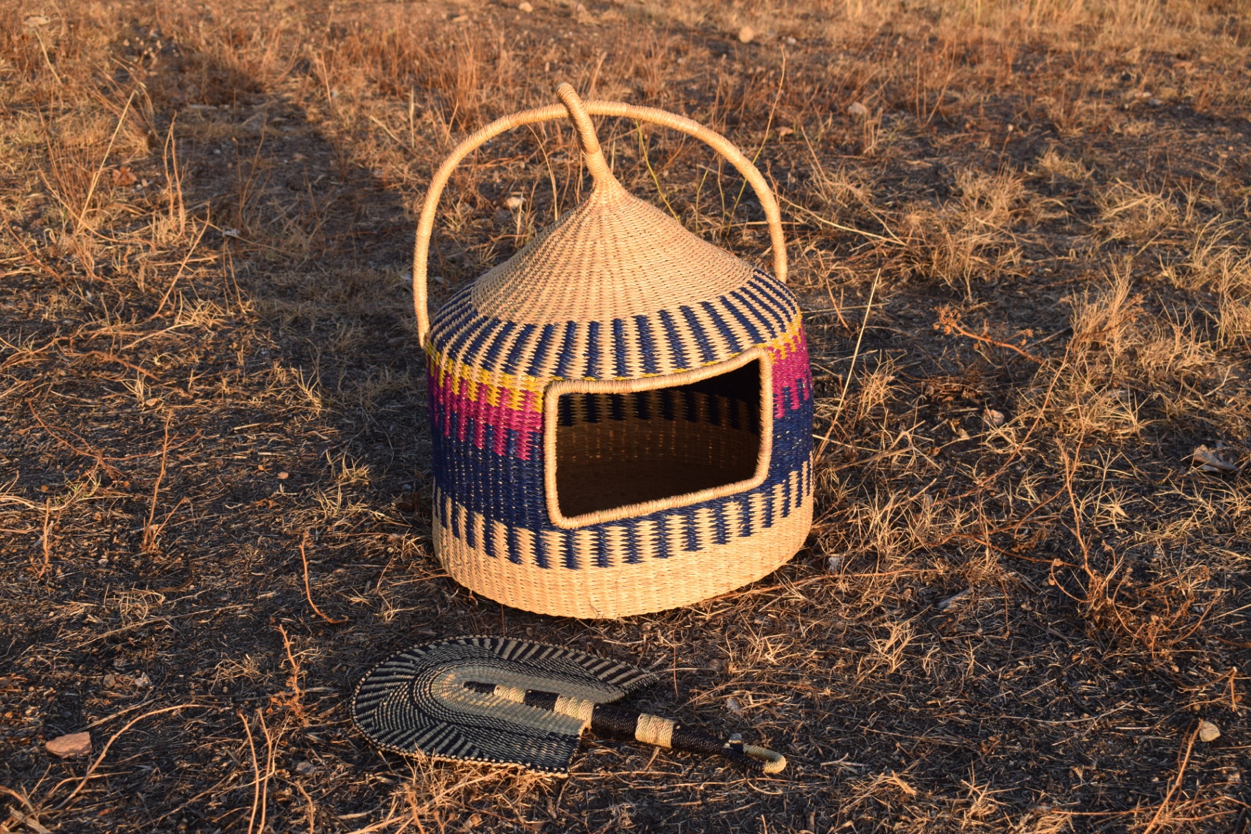 A finished Bolgatanga woven pet basket with blue, pink and yellow patterns set in a field.