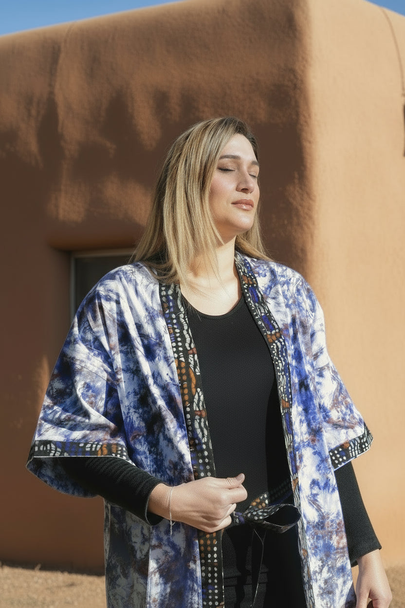 A woman standing outdoors wearing a multicolored long kimono with a geometric pattern, primarily in shades of blue, red, and white, with a border detail in a batik print over an Ankara base.