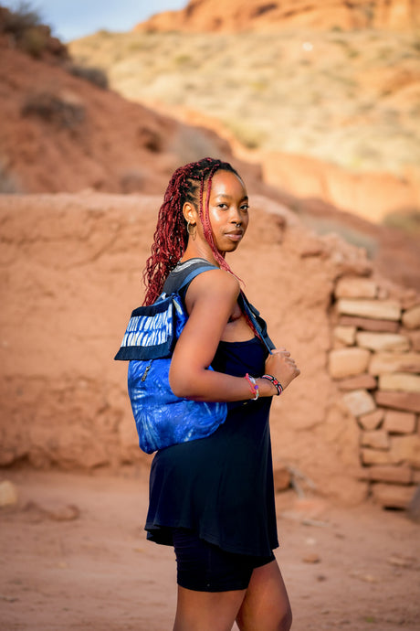 African girl with a blue tie dye backpack standing in an earthen environment.