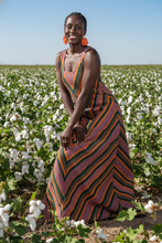 Load image into Gallery viewer, African woman wearing a handwoven smock dress in orange tones, standing in a cotton field
