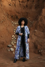 Load image into Gallery viewer, An African woman standing outdoors wearing a multicolored long kimono with a geometric pattern, primarily in shades of blue, red, and white, with a border detail in a batik print over an Ankara base.
