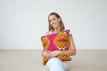 Load image into Gallery viewer, A person sitting outdoors with a cushion cover featuring a geometric pattern in pink, yellow, and burgundy colors with a yellow piping.

